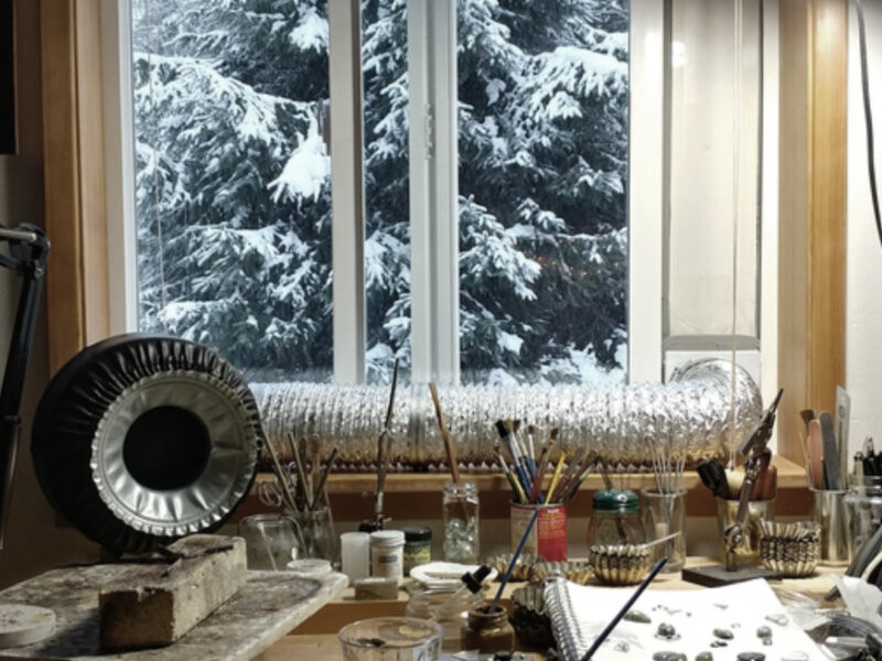A jeweler's bench scattered with tools and a soldering fume ventilation system in the background